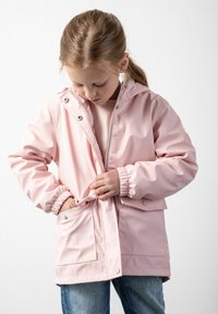 Jasnoróżowa wodoodporna kurtka z kapturem, z przodu na zamek, z zatrzaskami, elastycznymi mankietami i bocznymi kieszeniami. Modelka dostosowuje kurtkę.