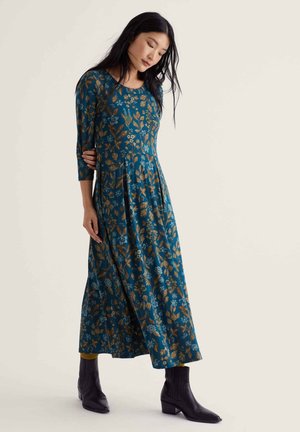 Femme en longue robe fleurie turquoise à manches trois-quarts, portant des bottines noires sur fond uni.
