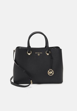 Sac à main Michael Kors noir avec ferrures dorées, double poignée, bandoulière amovible et un pendentif rond avec le logo MK sur fond blanc.