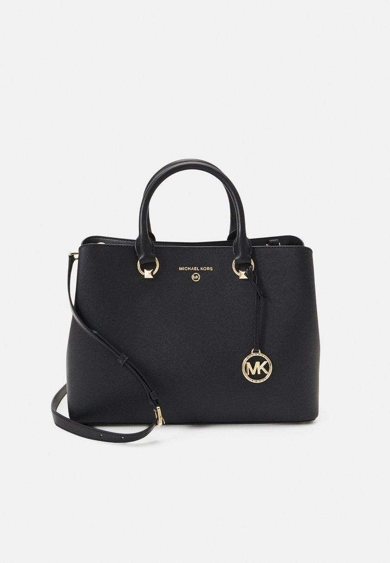 Sac à main Michael Kors noir avec ferrures dorées, double poignée, bandoulière amovible et un pendentif rond avec le logo MK sur fond blanc.
