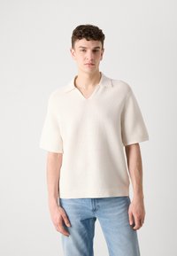 Kortermet, kremfarget polo-skjorte med v-hals, teksturvev og ribbestrikkede detaljer ved kragen og nederkanten. Sammen med lyseblå jeans.