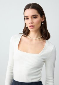 Top bianco a maniche lunghe con scollatura quadrata e bordi ondulati. Abbinato a una collana di perle e piccoli orecchini decorativi.