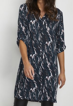 Kvinde iført en mørkeblå mønstret tunikakjole med lange ærmer, der er rullet op, stående mod en ensfarvet lys baggrund.