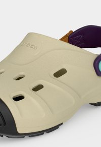 Μπεζ Crocs με οπές εξαερισμού, μαύρο κάλυμμα δακτύλων και μωβ-καφέ λουρί με λεπτομέρεια ανοιχτού μπλε κουμπιού.