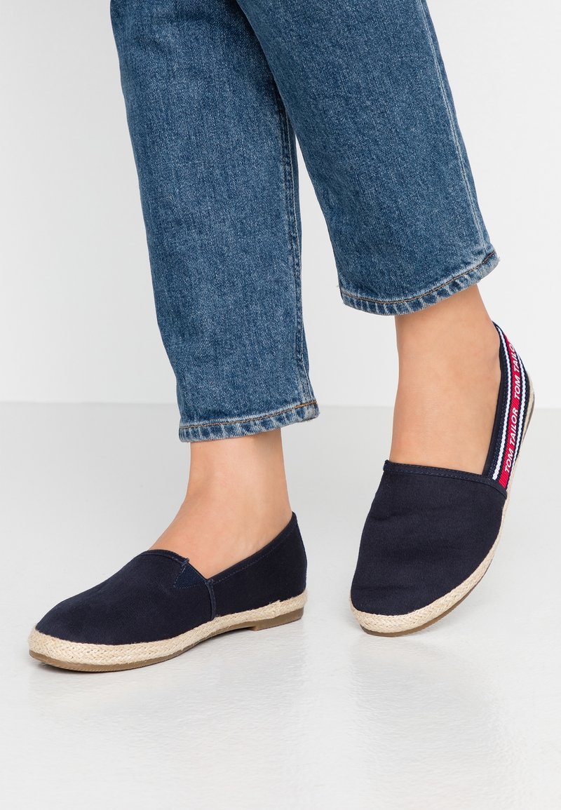 Personne portant des espadrilles bleu marine à enfiler avec des brides de talon blanches et rouges siglées et un jean bleu raccourci sur fond blanc.