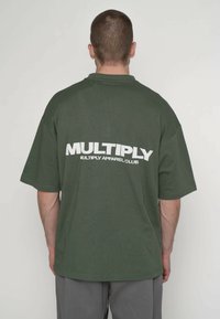 Oversized olivengrøn t-shirt med korte ærmer, med stort hvidt tekst på ryggen, der lyder "MULTIPLY APPAREL CLUB."