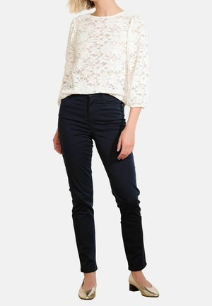 Kvinde iført en hvid blondebluse med trekvartærmer, mørke slim-fit bukser og metalliske sko med lav blokhæl, stående mod en hvid baggrund.