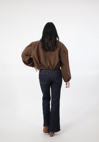 Brązowa skrócona bluza z elastycznym dołem, balonowymi rękawami, połączona z ciemnoniebieskimi, rozszerzanymi dżinsami. Modelka pokazana od tyłu, nosi brązowe szpilki.