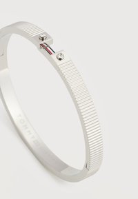 Βραχιόλι-μπανγκλ σε ασημί τόνο με ραβδώσεις, δύο μικρούς κρυστάλλους και λεπτομέρεια με ρίγες κόκκινου, λευκού και μπλε σε ένα λείο κούμπωμα.