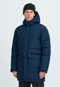 Tamno plavi zimski kaput s visokim ovratnikom, kapuljačom i dva prednja džepa. Izrađen od quilted materijala, ima ravni kroj i dugačke rukave.