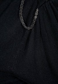 Svart texturerad tyg med kedjeaccent. Materialet verkar mjukt och har ett subtilt stickmönster. Kedjan har en blank finish.