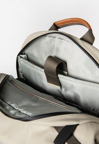 Beiger Rucksack mit einer glatten, wasserdichten Oberfläche, ausgestattet mit einem braunen Ledergriff, mehreren Fächern und einem weichen grauen Innenfutter.