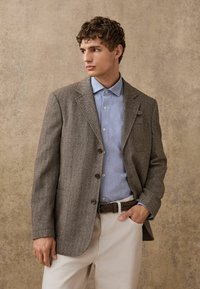 Herringbone-patterned brown blazer, paired with a light blue shirt and beige trousers. Features a single chest pocket and button closures.