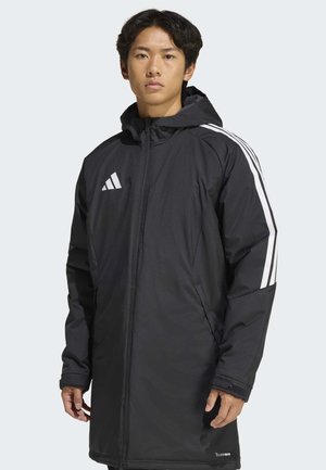 Jonge man draagt een lange zwarte Adidas-jas met capuchon en witte strepen op de mouwen, staand tegen een lichte achtergrond.