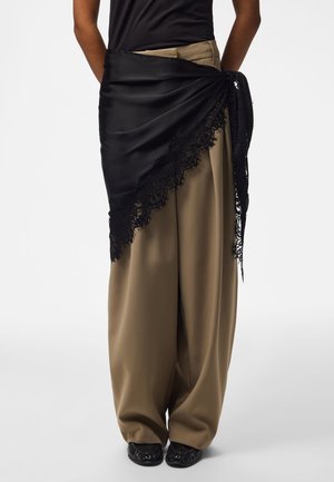Personne portant un pantalon large beige, un haut noir et une écharpe en satin noir avec une bordure en dentelle nouée autour de la taille, associée à des chaussures noires.
