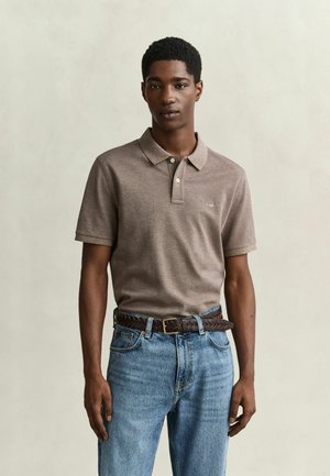Junger Mann trägt ein braunes kurzärmeliges Poloshirt, blaue Jeans und einen braunen geflochtenen Gürtel, steht vor einem einfarbigen hellen Hintergrund.