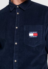 Navy corduroy overhemd met een button-down kraag, een bieszak met een "TOMMY JEANS" logo, en bruine knopen aan de voorkant.