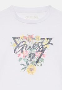 Weißes T-Shirt aus Bio-Baumwolle mit einem glitzernden Blumenmotiv innerhalb einer dreieckigen Umrandung und dem Schriftzug "Guess" in der Mitte.