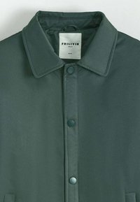 Veste vert foncé avec un collier, fermeture à bouton et une texture lisse. Étiquette visible avec le nom de la marque et les détails de la taille.
