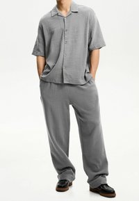 Graues Kurzarmhemd mit Kragen, geknöpft vorne; passende locker sitzende Hose. Strukturierter Stoff, entspanntes Design, kombiniert mit schwarzen Schuhen.