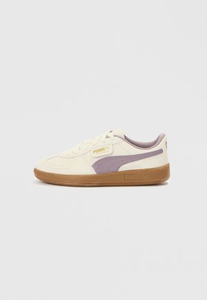 Baskets blanches Puma avec bande latérale violette et semelle en gomme, montrées de profil extérieur sur un fond clair uni.
