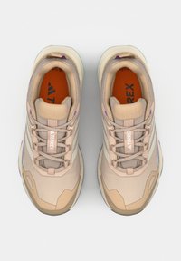 Pari beigenvärisiä Adidas Terrex lenkkareita, joissa on nauhat, pehmustetut kaulukset ja oranssit pohjalliset, kuvattu ylhäältä tasaisella taustalla.