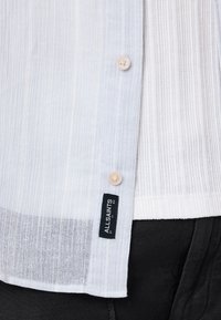 Svijetloplava teksturirana košulja s vertikalnim prugama, s patent zatvaračem i crnom oznakom branda, nosi se preko bijelog rebrastog topa i crnih hlača.