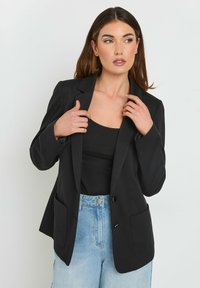 Blazer noir avec un bouton à l'avant, revers crantés et deux poches avant. Porté sur un top noir et associé à un jeans bleu clair.