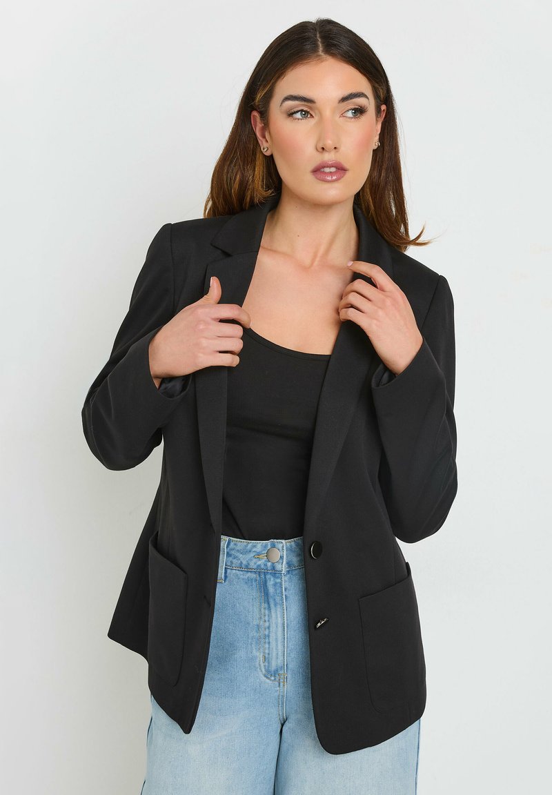 Blazer noir avec un bouton à l'avant, revers crantés et deux poches avant. Porté sur un top noir et associé à un jeans bleu clair.