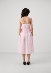 Rosa bomulls kjole med tynne stropper, tettsittende midje og rynket liv, som faller til midten av leggen. Båret med svarte sko med blokkheler.
