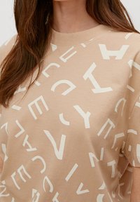 Beige t-shirt med store, kontrasterende hvide bogstaver i forskellige orienteringer. Rund halsudskæring og korte ærmer. Blødt bomulds materiale.