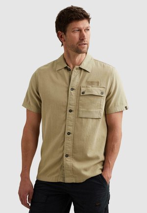Homme portant une chemise beige à manches courtes avec poche poitrine, regardant sur le côté, debout devant un fond uni gris clair.