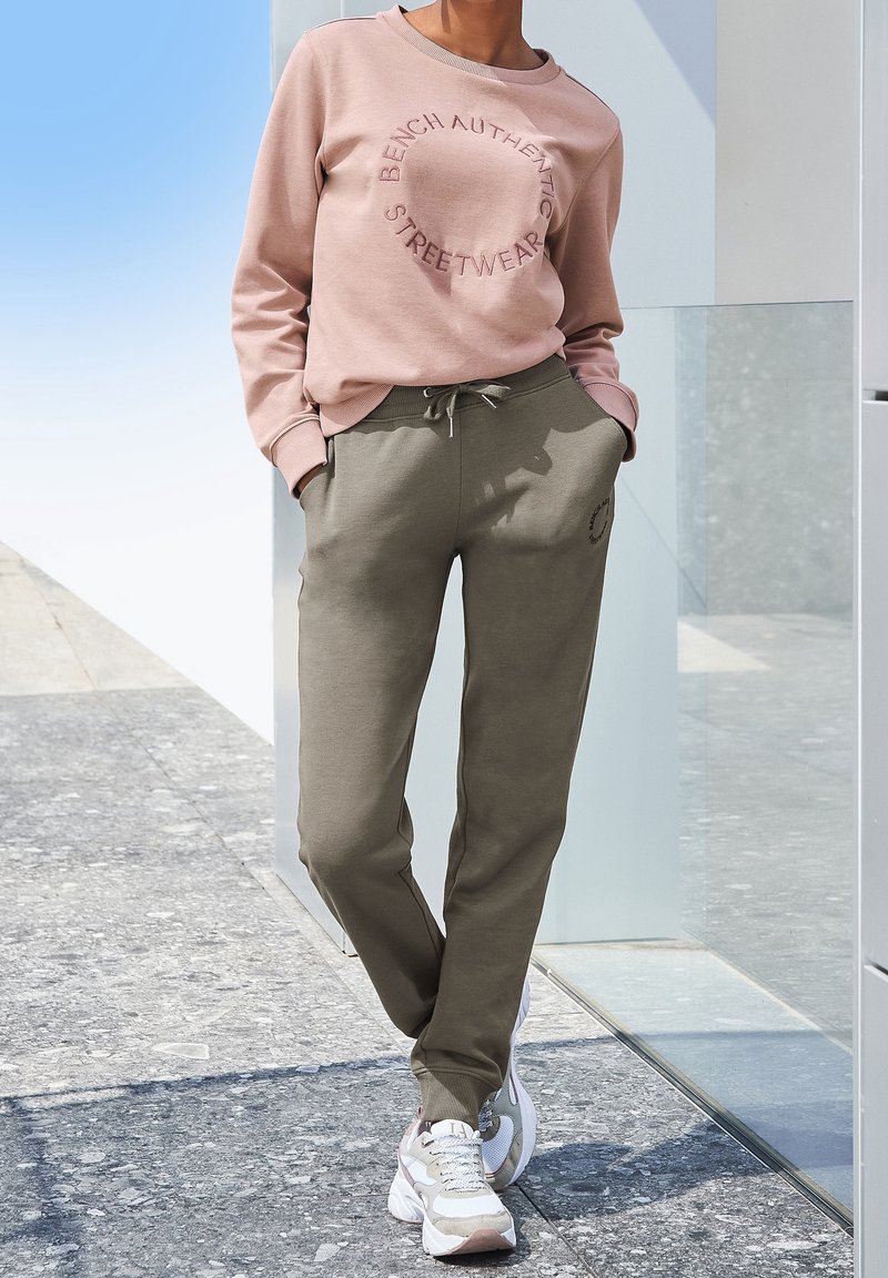 Personne portant un sweat-shirt rose avec le texte "Bench Authentic Streetwear" et un jogging vert olive, debout, les mains dans les poches et des baskets blanches.