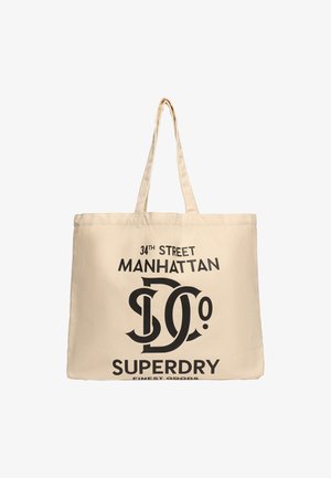 Beige canvas draagtas met twee handgrepen. Bevat vetgedrukte zwarte tekst: "34TH STREET MANHATTAN" en "SUPERDRY FINEST GOODS." Geen verdere patronen.