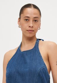 Denim halter topp i mørk blå med en tettsittende design, som har rene sømmer og åpen rygg. Jevn tekstur med et avslappet utseende.