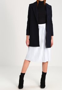 Granatowy długi płaszcz na czarnym golfie, zestawiony z białą plisowaną spódnicą midi i czarnymi botkami, o fakturze prążkowanej i pionowych liniach.