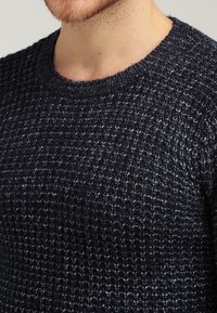 Niebieski sweter robiony na drutach z teksturowym wzorem i okrągłym dekoltem, wykonany z miękkiego materiału z widocznymi ściegami i ściągaczami.