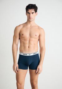 Tamno plave Nike bokserice s bijelim brendiranim pojasom, koje imaju prilagođeni dizajn i glatku teksturu. Model pozira s golim torzom.