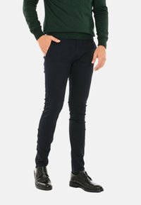 Pantaloni slim-fit blu navy realizzati in un misto di cotone elasticizzato, dotati di tasche laterali. Indossati con scarpe eleganti nere, che mostrano una texture liscia.