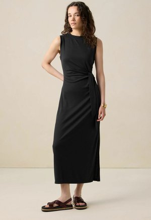 Robe maxi noire sans manches avec un design enroulé et une fente sur le côté. Tissu doux avec une texture lisse. Associée à des sandales à lanières marron.