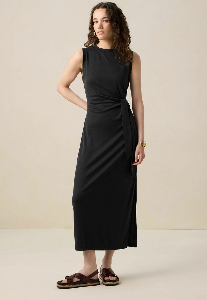 Robe maxi noire sans manches avec un design enroulé et une fente sur le côté. Tissu doux avec une texture lisse. Associée à des sandales à lanières marron.