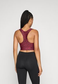 Kvinne med flettehår iført en rød og svart mønstret racerback sports-bh og svarte leggings, stående mot en enkel bakgrunn.