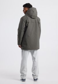 Giacca impermeabile verde scuro con cappuccio, caratterizzata da una linea dritta e una texture liscia. Indossata sopra pantaloni sportivi grigi, vista da dietro.