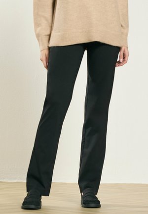 Persoon die zwarte rechte broek, zwarte instapschoenen en een beige trui met lange mouwen draagt, staande op een licht houten vloer.