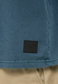 Blå bomullströja med en strukturerad yta, med en svart fyrkantig etikett prydd med "JACK & JONES" nära fållen.