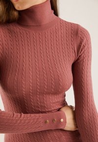 Gestrickter Rollkragenpullover in Rosafarbe, mit Rippendesign, Zopfmuster und Knopfdetails am Ärmelbündchen. Weiche Textur.