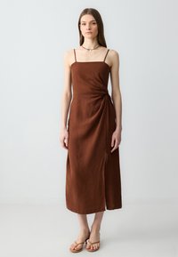 Abito midi in lino marrone con spalline sottili, caratterizzato da un dettaglio con nodo laterale e uno spacco. Indossato con sandali neutri su uno sfondo chiaro.