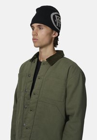 Veste en toile verte avec un col en velours côtelé, dotée de boutons sur le devant et de grandes poches. Cagoule noire avec un accent logo.