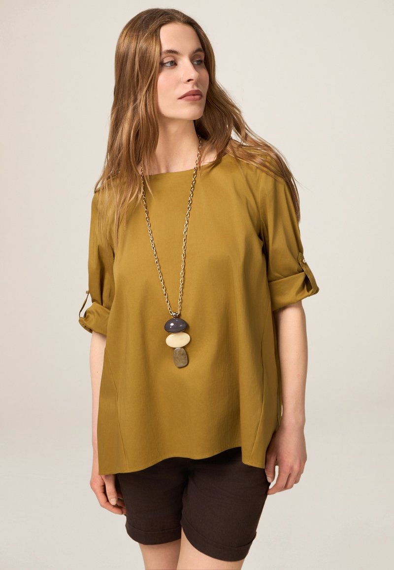 Senfgelbe Bluse mit hochgekrempelten Ärmeln, kombiniert mit braunen Shorts. Eine lange Halskette mit drei farbigen Steinen. Glatte Stofftextur.