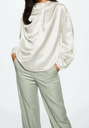 Femme portant un chemisier blanc en satin drapé à manches longues et un pantalon large taille haute gris clair, les mains dans les poches.